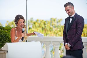 La mariée et le marié rient en prononçant leur discours de mariage. La mariée tient un microphone debout sur un podium blanc, tandis que le marié sourit à côté d'elle.
