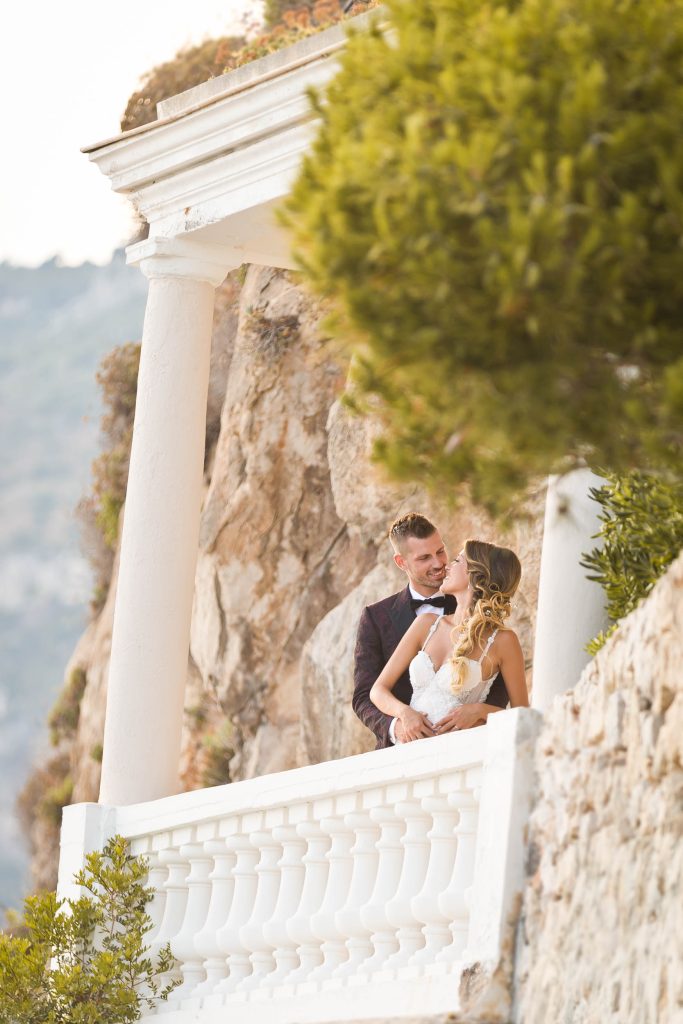 Un couple de jeunes mariés se tient dans les bras sur un balcon blanc surplombant la Côte d'Azur depuis un hôtel de luxe à flanc de falaise.