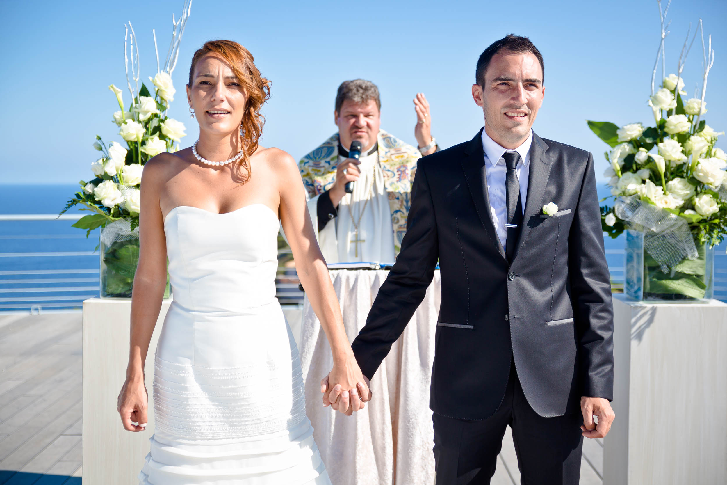 Un couple se tient la main en regardant vers l'avant lors de leur cérémonie de mariage avec leur cérémonian laïque et la mer derrière eux.